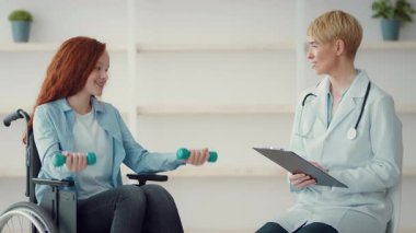 Bir terapist, tekerlekli sandalyedeki bir kıza, küçük ağırlıklar kullanarak bir güç antrenmanına katılması için rehberlik eder..