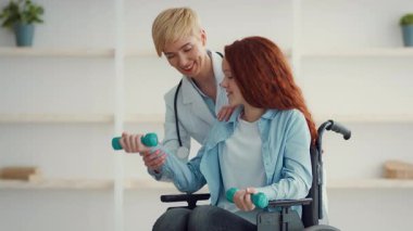Bir sağlık uzmanı, hafif ağırlık kullanarak fizik tedavi egzersizleri yapan tekerlekli sandalyedeki bir kızı destekler. Aydınlık ve misafirperver bir ortam. Rehabilitasyonu destekliyor..