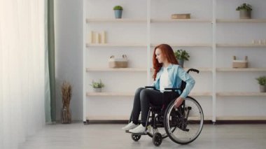 Kızıl saçlı kız tekerlekli sandalyede oturmuş, büyük pencerelerden dışarı bakıyor. Oda minimalist bir dekor içeriyor, sıcak ve davetkar bir atmosfer yaratıyor..