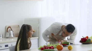 Bir baba ve kızı, her ikisi de gülümsüyor, güneşli bir mutfakta eğlenceli yemek pişirme faaliyetlerine girişiyor. Birlikte çeşitli taze sebzeler hazırlıyorlar, zamanlarının tadını çıkarıyorlar..