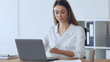 Genç bir kadın modern bir masada oturuyor. Etrafı parlak bir ofis ile çevriliyken dizüstü bilgisayarına odaklanmış. Gündüz saatlerinde iş ortamında meşgul ve üretken görünüyor..