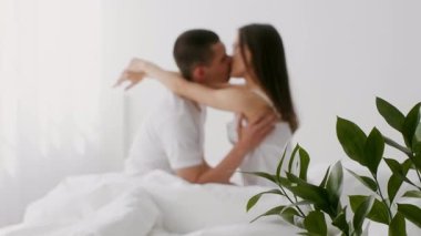 Genç bir çift, beyaz çarşaflar ve yeşilliklerle çevrili parlak bir yatak odasında şefkatle kucaklaşır. Sevgi ve sevgiyle vakit geçirirler..