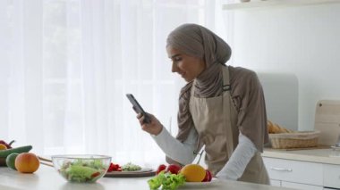 Orta Doğulu tesettürlü bir kadın açık bir mutfakta yemek hazırlamakla meşgul. Etrafı taze sebze ve meyvelerle çevriliyken telefonunu kontrol ediyor..