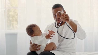 Afrikalı Amerikalı bir çocuk, bir bakıcının kucağında oturuyor ve eğlenceli bir etkileşime giriyor. İçerideki ortam aydınlık ve rahat, çocuk için besleyici bir atmosfer yaratıyor..