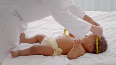 Afro-Amerikalı bir çocuk yumuşak bir yüzeyde yatarken bir yetişkin nazikçe sarı bir bantla kafasını ölçüyor. İçerideki sıcak ortam bu beslenme anına sıcak bir hava katıyor..