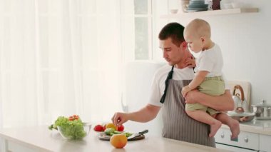 Bir baba, bebeğini tek koluyla tutar. Güneşli bir mutfakta taze meyve hazırlarken gülümser. Oda, tezgahında yeşil marul ve renkli meyvelerle parlak bir tasarım içeriyor..