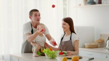 Bir bebek tezgahın üzerinde otururken ebeveynler parlak, rahat bir mutfakta taze sebzeler hazırlıyorlar. Aile birlikte iyi vakit geçirmeyi, bakım ve bağları geliştirmeyi seviyor..