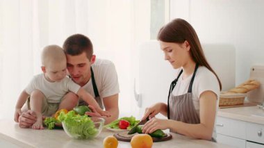 Ebeveynler sebze doğrayıp sağlıklı bir yemek hazırlarken bir bebek tezgahta oturur. Parlak mutfak, taze ürünlerle dolu, aile bağları yemek hazırlıkları sırasında sergileniyor..