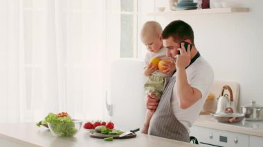 Bir baba, mutfak tezgahında taze sebzeler ve meyvelerle çevriliyken, küçük çocuğunu telefonda konuşurken tutar. Parlak, rahat bir ev ortamında beslenme anından hoşlanırlar..