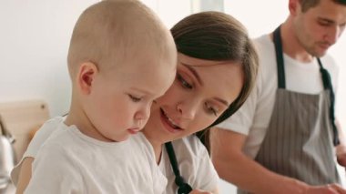 Bir anne sıcak bir yuva ortamında çocuğuna sevgi dolu bir şekilde bağlanır ve baba yakınlarda hazırlanırken çocuğun merakına odaklanır. Atmosfer özeni ve birlikteliği yansıtıyor..