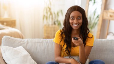 Ev eğlenceleri. Uzaktan kumandalı gülümseyen siyah kadın patlamış mısır yiyor ve oturma odasında kanepede televizyon izliyor. Afrikalı Amerikalı bir kadın ilginç bir televizyon programı seçiyor.