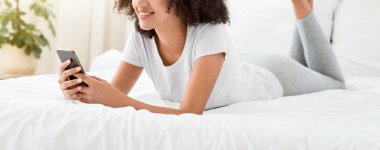 Tatil, hafta sonu, evde aletlerle ve sosyal ağlarla boş zaman. Mutlu bin yıllık Afro-Amerikan kadın ev kıyafetleri içinde yatağında uzanıyor ve modern yatak odasının içinde akıllı telefonuyla daktilo yazıyor.