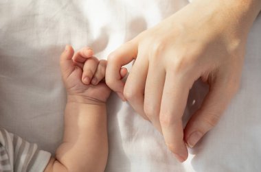 Anne Bebek Bağlantısı. Yeni doğmuş bir çocuk, annesinin elini tutarken yatakta yatar, küçük parmaklarıyla annesinin koluna dokunur, üst görünümü, kırpılmış görüntüsü, yakın plan.