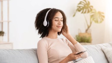 Kulaklıklı mutlu komik bayan rahat bir koltukta oturur, yeni pop müzik dinler, gözleri kapalı şarkıdan zevk alır, evdeki rahat oturma odasında dinlenir. Genç Afro-Amerikan kadın telefonu tutuyor.