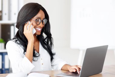 Ofis kıyafetleri içinde, iş yerinde cep telefonuyla konuşan, dost canlısı, genç, siyah bir kadın. Çekici Afrikalı Amerikalı girişimci müşterisiyle iş telefonu görüşmesi yapıyor