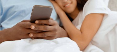 Evde aletlerle modern tatil, haberleri okumak ve yatakta internette sörf yapmak. Gülümseyen bin yıllık Afro-Amerikan kadın ve erkek sarılır ve akıllı telefona bakar, yatak odasında, panorama 'da, fotokopi odasında, kırpılmış