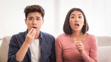TV 'de korku filmi seyreden Koreli çift, evdeki oturma odasındaki koltukta oturmuş patlamış mısır yiyor. Çinli Aile, Hafta sonu için Televizyon, Korku Filmleri İzliyor