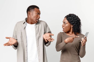 Gadget ve sosyal medya bağımlılığı, ilişkilerde hile. Siyah kadın erkek arkadaşının cep telefonunu kontrol ediyor kışkırtıcı mesajlar gösteriyor ve adama bağırıyor, gri stüdyo geçmişi var.