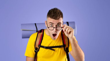 Şok olmuş genç bir adam, turist ekipmanlarıyla kameraya bakıyor ağzı açık, mavi stüdyo geçmişi var. Gözlüklere dokunup şaşkınlığını ifade eden tuhaf bir sırt çantası.