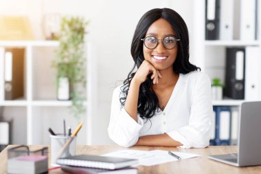 Modern ofiste dizüstü bilgisayarla masasında oturan Afro-Amerikan bir iş kadını. Siyahi kadın asistan kameraya bakıyor ve gülümsüyor. Bilgisayarı internet iletişimi için kullanıyor.