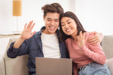 Neşeli Asyalı çift dizüstü bilgisayarda video görüşmesi yapıyor koltukta oturuyor, el sallıyor. Çinli Aile Uzaktan Dostları Çevrimiçi, Uzaktan İletişim Konsepti
