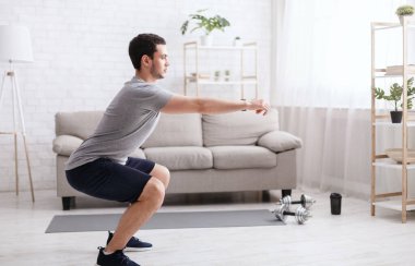 Çalışmak için evde tek başına kal. Oturma odasında çömelme egzersizi yapan genç adam. Evin içinde, boş alanda.