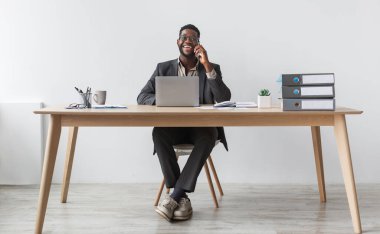 Cheerful black businessman in formal suit talking on smartphone, having conversation, sitting at table with laptop against white studio wall, copy space. Remote communication concept