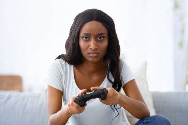 Evde oyalanalım. Oyun çubuğuyla kanepede video oyunu oynayan ciddi bir siyahi kadın. Odaklanmış Afrikalı Amerikalı kadın tecrit sırasında bilgisayar oyunlarıyla kendini eğlendiriyor.