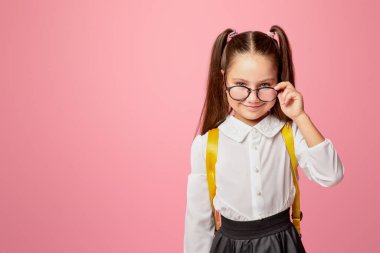 Okulun ilk günü. Üniformalı neşeli kız gözlüklerini çıkarıp kameraya bakıyor, pembe arka planda izole edilmiş, stüdyo çekimlerinde.