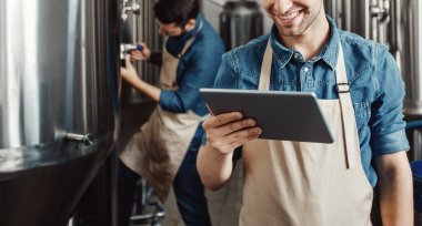 Bira fabrikasında bira üretim süreci. Bira fabrikasında bira üreten ve dijital tablet kullanan bir adam. Önlüklü neşeli genç yakışıklı adam büyük su ısıtıcısının verilerini el yapımı içecekle kontrol ediyor.