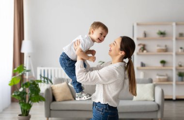 Emotional young caucasian mother raising laughing adorable toddler baby son, holding kid pretending flying, having fun and playing together in light living room, side view