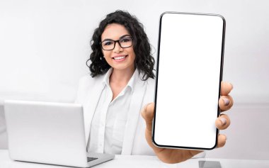 Female doctor showing big smartphone with blank screen, sitting in office at workplace, closeup, mockup. Professional physician advertising medical mobile application or website
