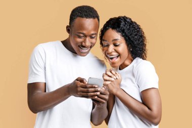 Heyecanlı siyah çift cep telefonu ekranına bakıyor, yeni bir uygulama kullanıyor, sarı stüdyo geçmişi var. Neşeli Afrikalı Amerikalı erkek ve kadın akıllı telefon kullanıyor, eğlence konsepti.