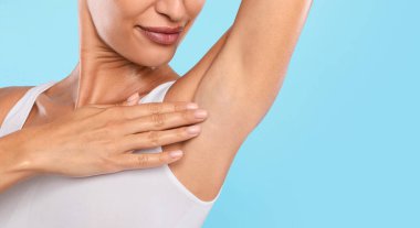 Mavi stüdyo arka planında izole edilmiş koltukaltını koklayan, elini kaldırıp Axilla 'ya dokunan mutlu kadının yakın plan görüntüsü. Ücretsiz fotokopi alanı, koltuk altı deodorantı veya epilasyon reklamı