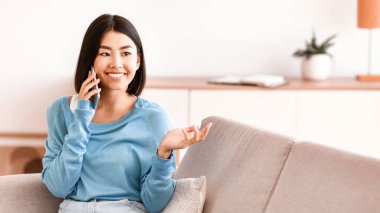Arkadaş 'la hoş sohbetler. Çekici gülümseyen Asyalı kadın cep telefonuyla konuşuyor, oturma odasındaki kanepede oturuyor. İletişim Konsepti. Bayan cep telefonuyla konuşuyor.