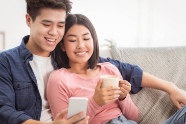 Hafta sonları evde koltukta oturan akıllı telefon kullanan Koreli çift. Cep telefonu kullanıcıları interneti tarıyor, Gadget Yaşam Tarzı, Mobil Uygulama Kavramı. Boş Boşluk