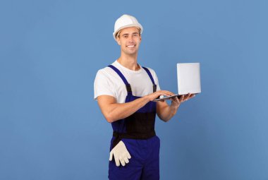 Mavi stüdyo arka planında dizüstü bilgisayar kullanan, tulumlu ve kasklı gülümseyen genç adam, çevrimiçi yenileme için araç ve malzeme sipariş eden yakışıklı erkek işçi, kopyalama alanı