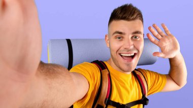 Kamp vitesleriyle milenyum adamı selfie çekiyor ve mavi stüdyo arka planında akıllı telefon kamerasına el sallıyor. Neşeli genç yürüyüşçü turist gezisinden önce havalı bir fotoğrafını çekiyor.