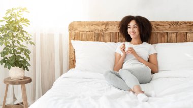 Hafta sonu, günaydın, pozitif ruh hali ve canlılık. Mutlu Afro-Amerikan kadın yatakta oturuyor, elinde bir fincan sıcak içecekle ve rahat bir sabah geçiriyor modern yatak odasının içinde, boş bir yerde.