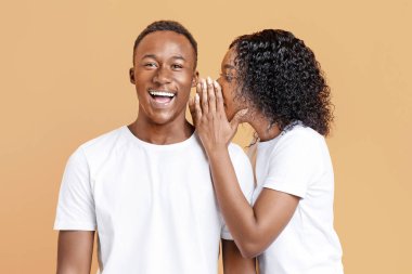 African-american young woman sharing secret or whispering gossips into her boyfriends ear, yellow studio background. Black lady whispering words of love to her excited and happy man