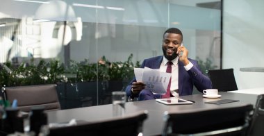 Takım elbiseli zengin siyahi girişimci asistanı veya iş ortağıyla telefon görüşmesi yapıyor, dijital tabletle masada oturuyor, belgelere bakıyor, fotokopi alanı olan panoramaya bakıyor.
