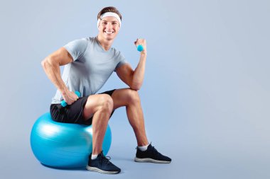 Spor giyimli, spor kıyafetli, spor topu üzerinde oturan ve Violet stüdyosunun arka planında dambılları kaldıran genç bir adamın tam boy portresi. Daha sağlıklı yaşam konsepti için egzersiz