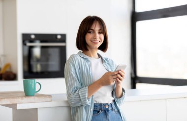 Gülümseyen, akıllı telefon kullanan genç bir bayan mutfaktaki kahvenin tadını çıkarıyor, masanın yanında duruyor ve kameraya gülümsüyor, boşluğu kopyalıyor.