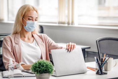 Antiseptikle birlikte çalışma odasındaki modern işyeri. Maskeli güzel bir iş kadını Coronavirus karantinası sırasında laptopuyla masada oturuyor.