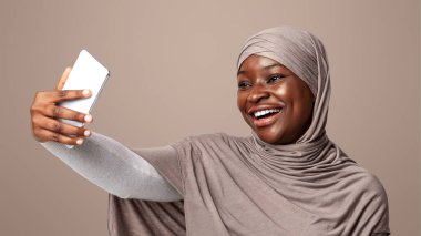 Neşeli siyahi kadın tesettürde selfie çekiyor, neşeli Afrikalı İslamcı kadın kendi portresini yapıyor, kamera karşısında poz veriyor ve gülümsüyor, ışık stüdyosunun arka planında duruyor.