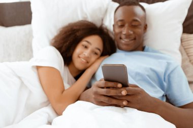 Aşk, ilişki, teknoloji, hafta sonu ve birlikte tatilin tadını çıkarın. Rahat yatak odasında gülümseyen Afro-Amerikan erkek ve kadın sabah içteki telefona bakan adam, uzay boşluğunu kopyalayan...