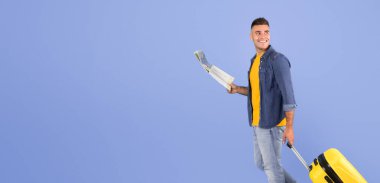 Mavi stüdyo arka planında yürüyen bavulu ve haritası olan genç bir adamın tam boy portresi. Kopya alanı. Yolculuk güzergahını çizen bagajlı neşeli çekici bir gezgin.