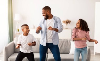 Eğlence Faaliyetleri Konsepti. Neşeli Afrikalı Amerikalı baba çocuklarıyla dans edip müzik eşliğinde dans ediyor. Kız, erkek ve baba evde, oturma odasında aileleriyle eğleniyor.