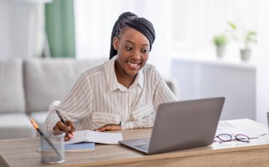 Genç siyahi bayan evde online eğitim alıyor, dizüstü bilgisayar kullanıyor ve notlar alıyor, boşluğu kopyalıyor. Gülümseyen Afrikalı Amerikalı kadın çevrimiçi konferansa katılıyor, görevler yazıyor, oturma odası içleri...