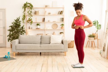 Kilo kaybı konsepti. Üzgün siyah kız, şişman göbeğine dokunuyor evde zayıflayan pulların üzerinde duruyor. Boş Boşluk